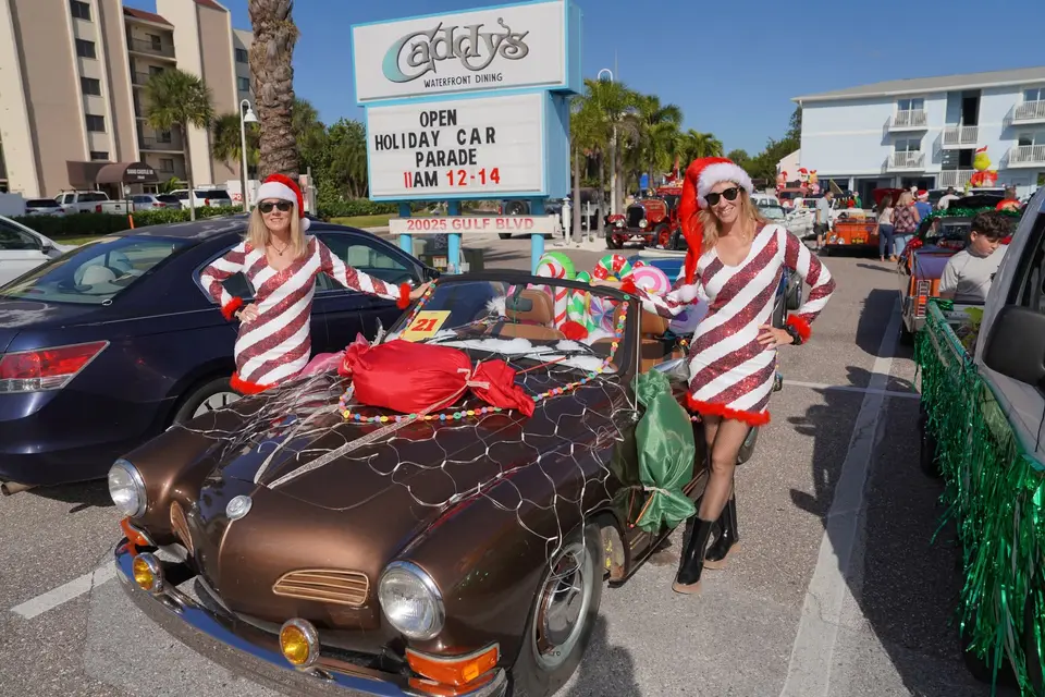 Two women in candy-cane dresses with a gift-wrapped Karmann Ghia under Caddy's Holiday Car Parade sign