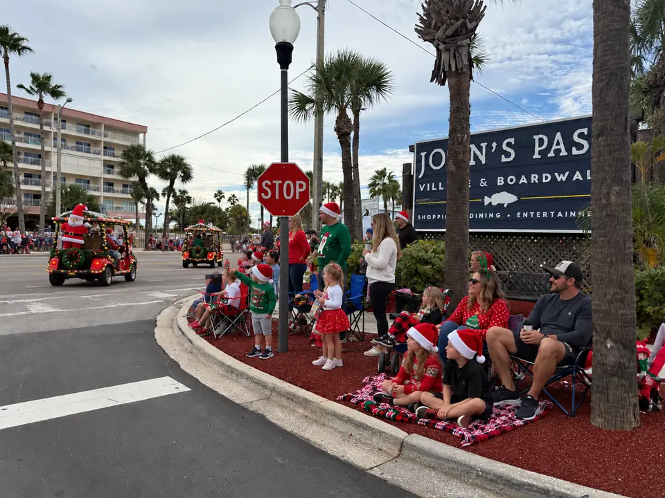 Families in Santa hats sitting on the curb by the John's Pass Village sign, waving as decorated golf carts roll by